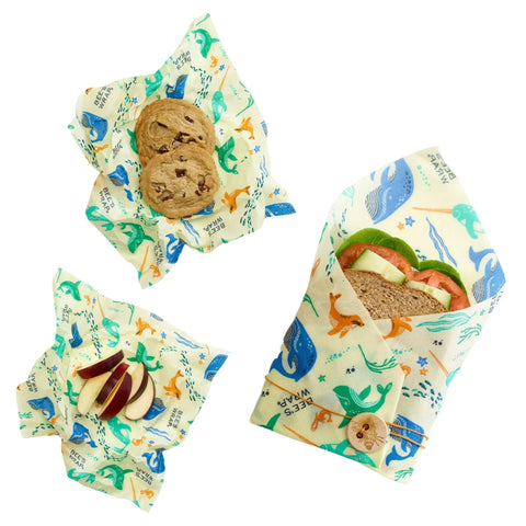 The Lunch Pack by Bees Wrap features three reusable beeswax food wraps with colorful dinosaur prints, holding cookies, apple slices, and a lettuce-tomato sandwich. One compostable wrap with a wooden button is shown on a white background.