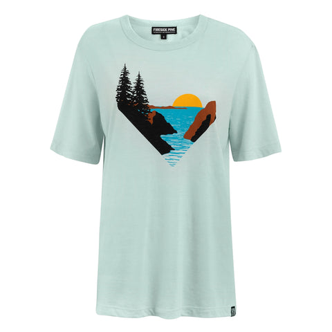 The Rocky Mountain Juniper - Sky Blue T-shirt by Fireside Pine features a soft combed cotton oversize fit and a central Pacific Ocean coastline graphic with pine trees, rocks, water, and an orange sunset for a striking nature scene.