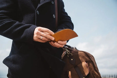 A person in a dark coat holds the Rogue Industries Rogue Front Pocket Wallet in Ballglove and a brown bag outdoors beneath a cloudy sky.