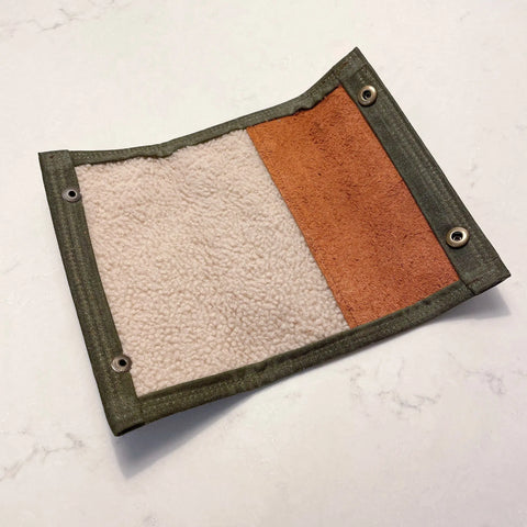 The Rogue River Fly Fishing Wallet by Rogue Industries is a rectangular swatch with cream fleece on one side, brown suede on the other, green fabric edges, and metal grommets along two sides, shown lying flat on a light surface.