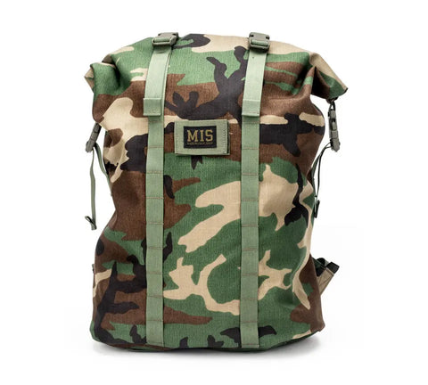 The MIS Roll Up Backpack - Woodland Camo features Cordura Nylon in a camouflage pattern, green straps, roll-top closure, heavy-duty YKK zipper, and a rectangular MIS patch on the front. Photographed against a white background.