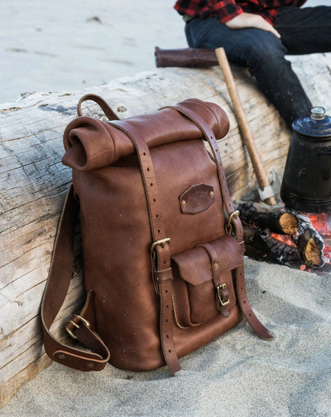The Urban Southern Rolltop Backpack, designed for weather resistance, rests on sandy ground near a campfire and gear, with a person in a red plaid shirt partly visible behind driftwood.