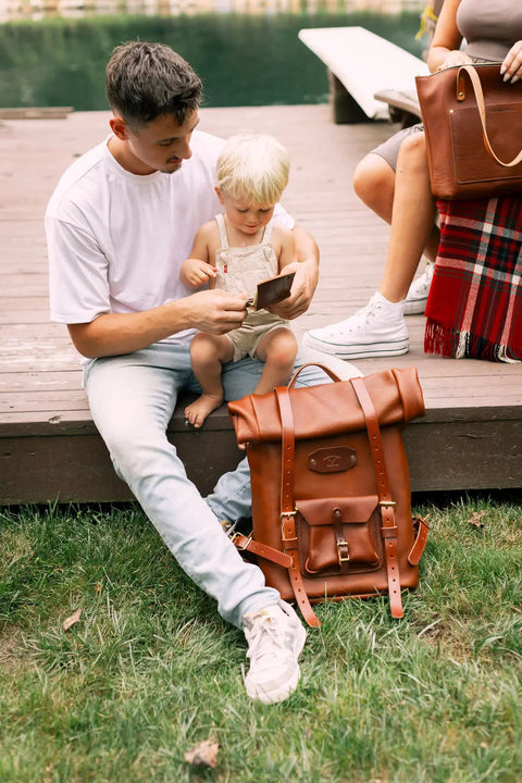 A man sits on wooden steps with a child on his lap, while an Urban Southern Rolltop Backpack rests on the grass nearby. Another person, carrying a brown leather bag, is partially visible.