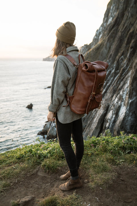 Wearing a beanie, gray shirt, black pants, and brown boots, a person with the Urban Southern Rolltop Backpack stands on a grassy cliff edge, gazing over the ocean as rocky cliffs and vast water create a dramatic backdrop.