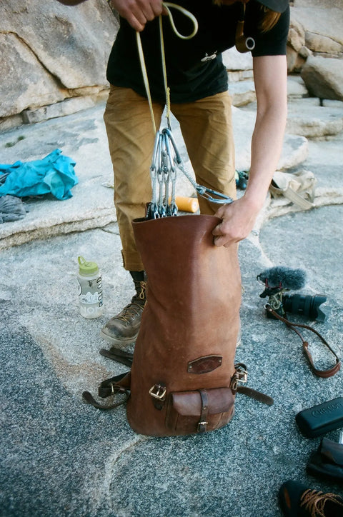 On a rocky surface, someone packs climbing gear into the Urban Southern Rolltop Backpack, with a water bottle, camera, microphone, and other essentials scattered around.
