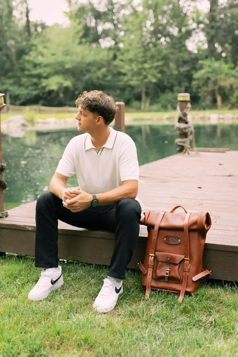 A man in a white polo and black pants sits on a wooden dock by a pond, looking to the side. Next to him on the grass is the Urban Southern Rolltop Backpack, weather-resistant leather, surrounded by trees and greenery.