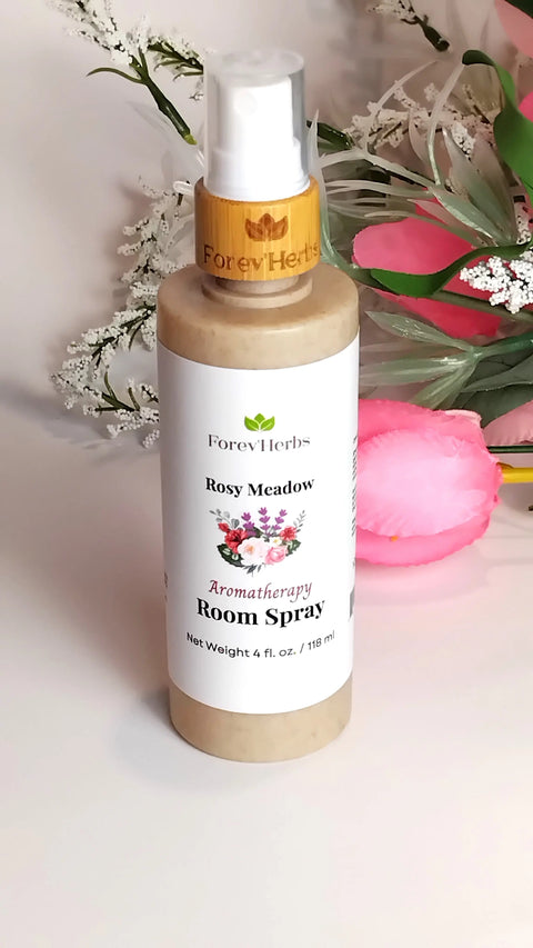 A bottle of Forev'Herbs Room Spray rests on a white surface, surrounded by artificial pink flowers and white floral accents.