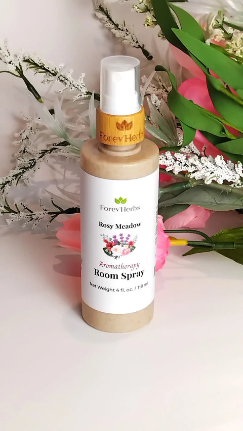 A Forev'Herbs Room Spray bottle stands upright among pink and white flowers and green leaves, featuring an eco-friendly design with a white label accented in green and pink.