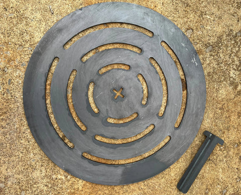 The Ohio Flame Round Log Grate (Made in USA) is a black steel plate with circular slits and an X-shaped center, designed for fire pits. Shown on concrete beside a black handle with a T-shaped top.