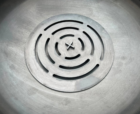 A close-up of an Ohio Flame Round Log Grate (Made in USA), featuring concentric circular and curved slots, secured by a central screw and set in a smooth metallic surface—crafted from quality American steel.