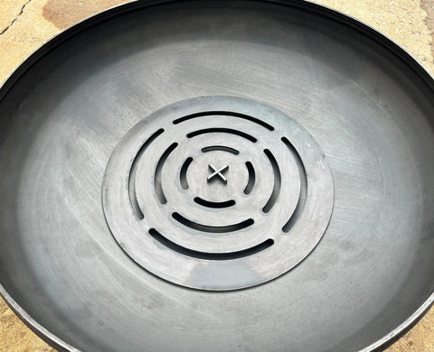 A close-up of the Ohio Flame Round Log Grate (Made in USA), a large, round black metal fire pit accessory with circular grooves and central slots, seen on a concrete surface.