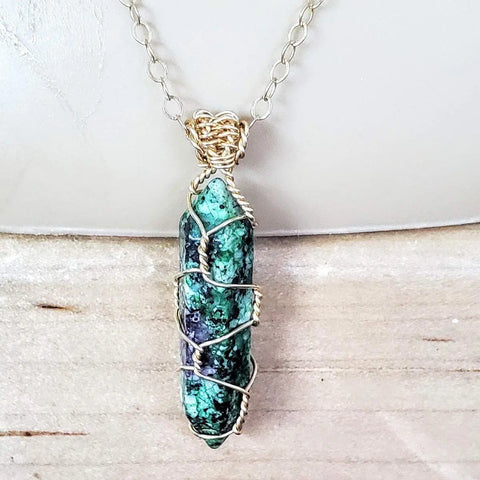 The Alexa Martha Designs Gold Wire Wrapped Caged in Ruby in Zoisite Pointed Crystal Necklace features a crystal pendant wrapped in gold wire on a delicate gold chain, shown against a light beige background.