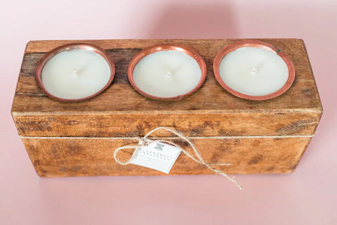 The Shanti + Tula Rustic Wooden Sugar Mold Candle features three clean-burning soy wax candles in copper holders, arranged in a handmade 3-hole dough bowl with a twine and tag, set against a pale pink background.