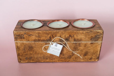 The Shanti + Tula Rustic Wooden Sugar Mold Candle features three clean-burning soy wax candles in copper tins, arranged in a handmade 3-hole dough bowl and tied with twine, set against a pale pink background.