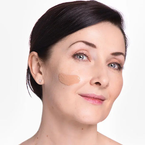 A woman with fair skin and short dark hair smiles slightly, displaying a swatch of Open Formula SPF 50 Tinted Moisturizer in beige-bronze on her cheek, set against a plain white background.