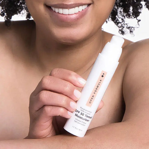 A smiling person with curly hair holds the Open Formula SPF 50 Tinted Moisturizer near their face, showcasing its broad spectrum protection to the camera.
