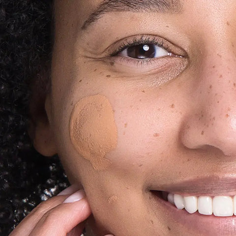 A close-up of a smiling person with curly hair wearing Open Formula SPF 50 Tinted Moisturizer on their cheek. Their teeth show as they pose with a hand by their chin.