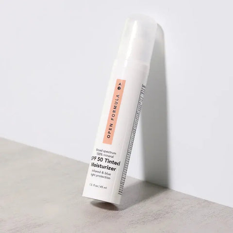 A white cylindrical bottle of Open Formula SPF 50 Tinted Moisturizer with a bronze tint and clear cap stands upright on a light surface against a textured, light gray background. Broad spectrum SPF 50 helps prevent premature aging.