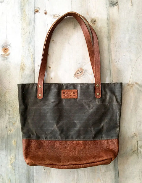 The Saco River Tote Bag by Rogue Industries, made in Maine with waxed canvas, full-grain leather handles and bottom, and a front leather patch, is shown on a light wooden surface.