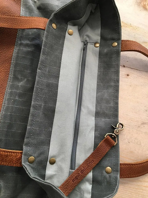 A close-up of the Rogue Industries Saco River Tote Bag shows brown full-grain leather straps, brass rivets, a central zipper, and an interior key leash stamped with Sage Industries. The waxed canvas bag rests on a wooden surface.