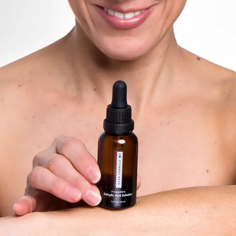 A person applies Open Formula Salicylic Acid Exfoliant from a brown dropper bottle to their bare shoulder, smiling slightly as they target clogged pores and acne scars.