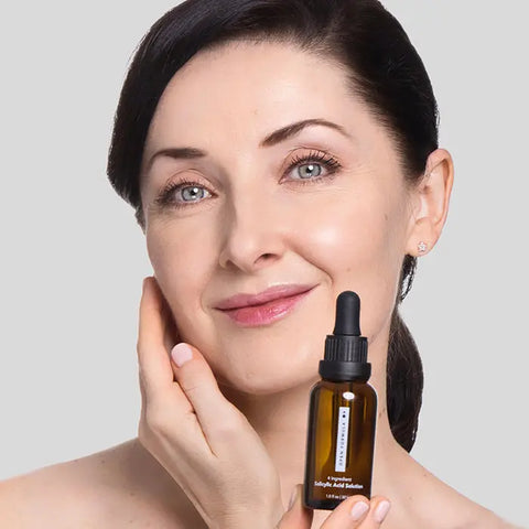 A woman with fair skin and dark hair smiles, gently touching her face while holding an Open Formula Salicylic Acid Exfoliant dropper bottle for clogged pores, against a light gray background.