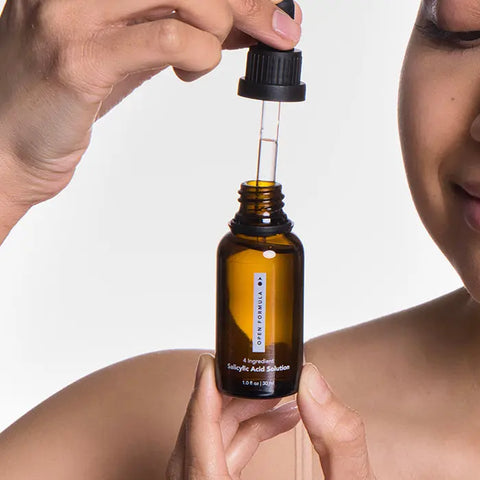 A person holds a dropper above an amber bottle labeled Salicylic Acid Exfoliant by Open Formula, ready to dispense the liquid—ideal for clearing blackheads and clogged pores—with part of their face and shoulder visible.