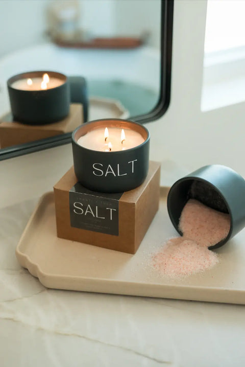 A black SALT Candle by Shanti + Tula, with two flames, sits by a tipped container spilling pink salt onto a beige tray, symbolizing hope and moving forward; set before a bathroom mirror and bathtub. Made in the USA.
