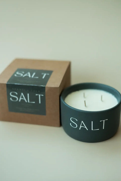 The Salt Candle – Faith-Based Gift by Shanti + Tula sits beside its matching brown box, both on a plain beige background. This black candle, with four unlit wicks, symbolizes hope and moving forward. Made in the USA.