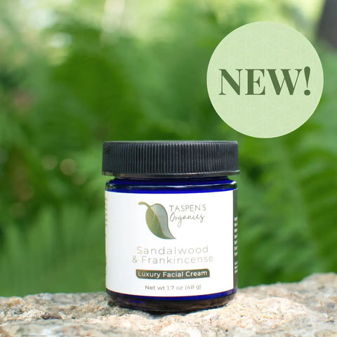 A jar of Taspens Organics Sandalwood & Frankincense Luxury Facial Cream sits outdoors on a stone surface, surrounded by green foliage, with a NEW! circle in the upper right corner.