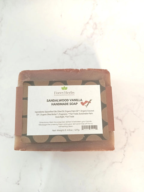 A brown Forev'Herbs Sandalwood Vanilla Handmade Soap bar, featuring a masculine scent, rests on a white surface. The label displays ingredients and the product weight (4.5oz / 127g).