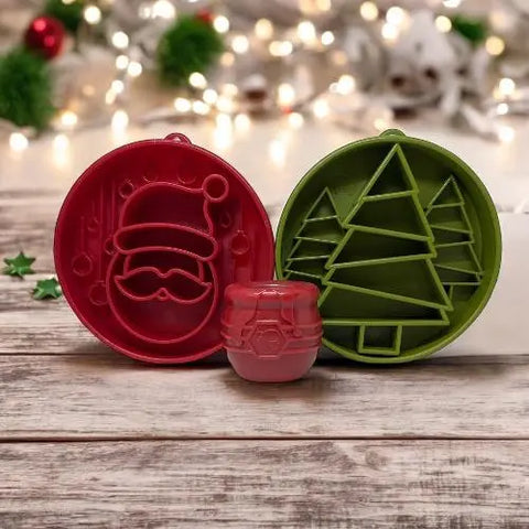 Red and green holiday cookie cutters shaped like Santas face and a Christmas tree rest on a wooden surface next to a red cookie and the SodaPup Santa Design EBowl Enrichment Slow Feeder Bowl for Dogs, with glowing decorations in the background.