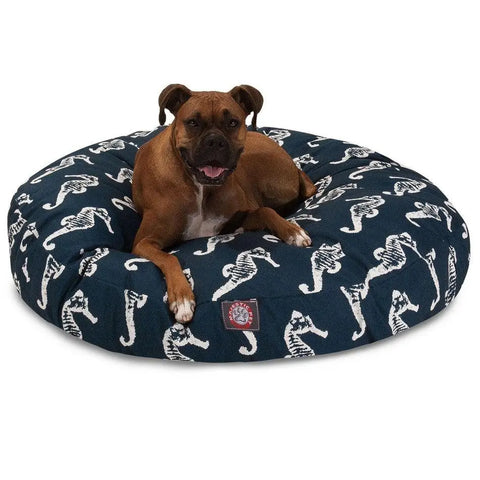 A happy brown dog lies on the Majestic Pet Sea Horse Round Dog Bed, featuring a removable, washable cover in navy blue with white seahorse patterns. The pups mouth is open and its ears are perked up.