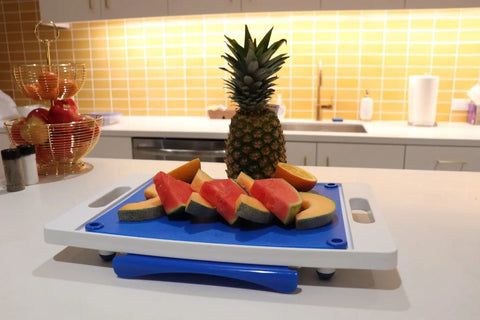 A Karving King Set of 2 Dripless Cutting Boards rests on a white kitchen counter with sliced watermelon and cantaloupe. A pineapple is behind the board, and apples fill a basket against a yellow tiled backsplash.