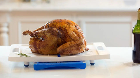 A whole roasted turkey sits on a Karving King Set of 2 Dripless Cutting Boards 2 in 1 System with handles, on a kitchen counter. A sprig of rosemary is beside the turkey, and part of a bottle appears to the right.