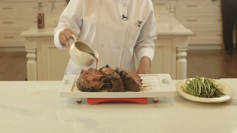 A chef uses the Karving King Set of 2 Dripless Cutting Boards 2 in 1 System to pour sauce over roasted meat, with asparagus and green beans on the side, in a bright kitchen with white cabinets.
