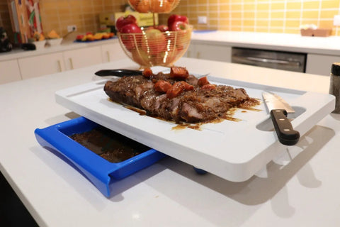 A cooked steak with sauce sits on the Karving King Set of 2 Dripless Cutting Boards 2 in 1 System, featuring a blue tray for catching juices, on a kitchen counter. A black knife and fruit are also visible in the background.