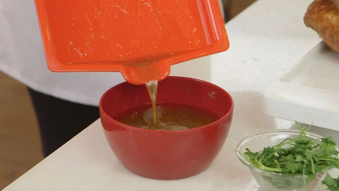 Someone pours liquid from a Karving King Set of 2 Dripless Cutting Boards 2 in 1 System into a red bowl on the counter, with a glass bowl of fresh greens and part of a cooked dish on a white tray nearby.