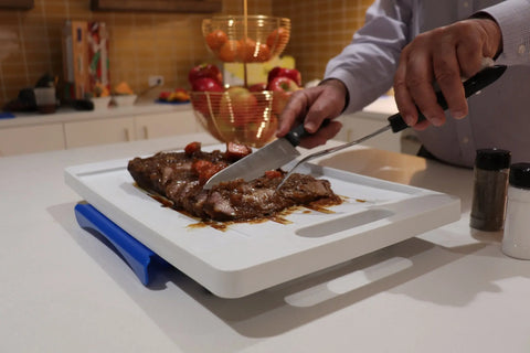 In a kitchen, someone slices cooked meat with a knife and fork on the Karving King Set of 2 Dripless Cutting Boards 2 in 1 System. A fruit basket with apples and oranges sits nearby, along with salt and pepper shakers.