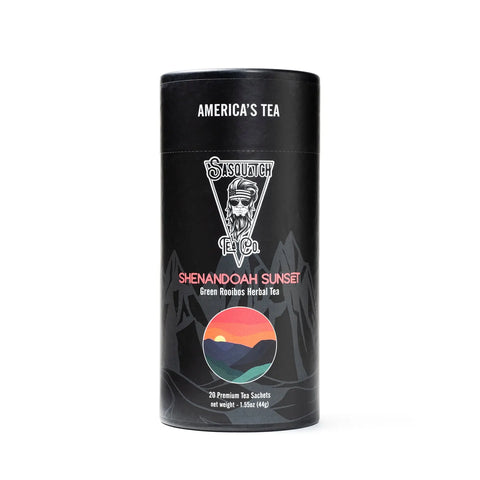 A black cylindrical tea container from Sasquatch Tea Co, labeled Shenandoah Sunset (Green Rooibos Herbal Tea), features colorful sunset and mountain graphics with Americas Tea at the top. This caffeine-free blend offers a unique herbal experience.