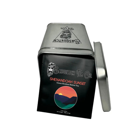 A silver tin tipped on its side with the lid off reveals a bag labeled Sasquatch Tea Cos Shenandoah Sunset (Green Rooibos Herbal Tea), a caffeine-free tea with a vibrant mountain illustration.