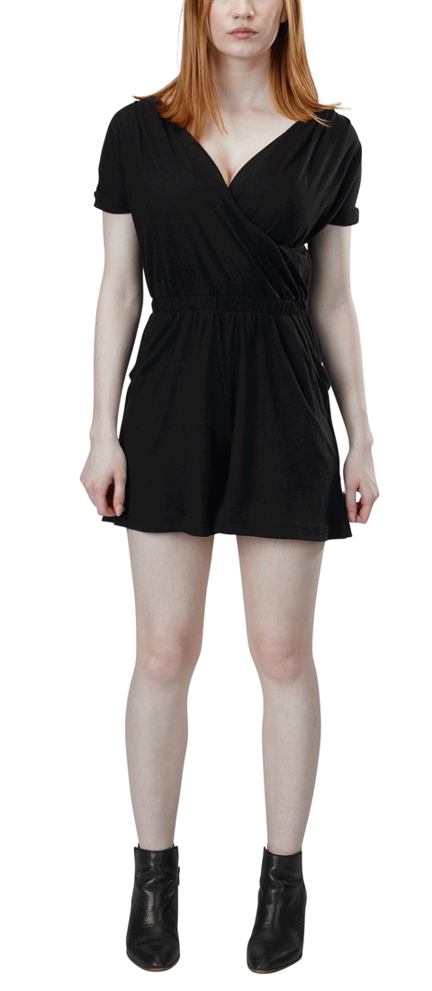 A woman with straight, shoulder-length hair wears the Spiritex Short Sleeve Surplice Romper in black, made from organic cotton/Tencel. The romper features a V-neck and short sleeves. She pairs it with black ankle boots and stands facing forward, arms relaxed.