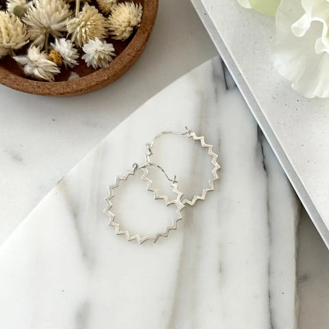 A pair of SILVER POINTE HOOPS by Sarah Cecelia sits on a white marble surface beside a bowl of dried flowers.