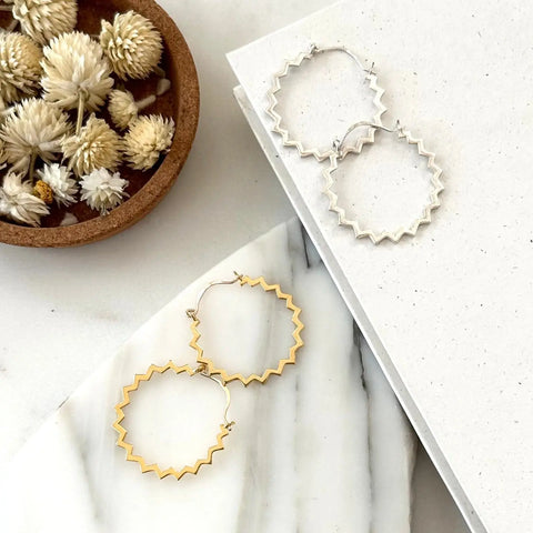 The Sarah Cecelia SILVER POINTE HOOPS and gold zigzag hoops are displayed on a white marble and textured surface beside a small bowl of dried white flowers.