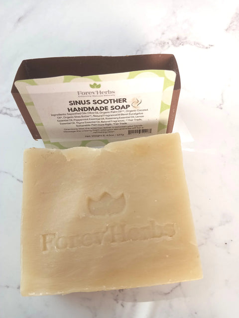A beige bar of Forev'Herbs Sinus Soother Handmade Soap sits on a white surface, with a brown and green label showing ingredients and brand details behind it.