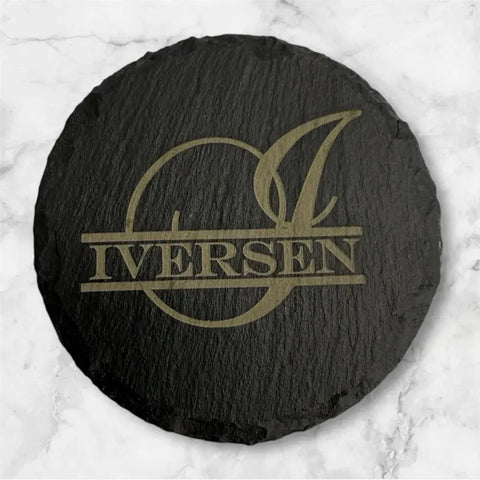 A round black slate coaster from the Slate Coaster Set of 6 by B. James Design & Woodwork features a large stylized I over a circle with IVERSEN in bold and rests on a white marble surface.
