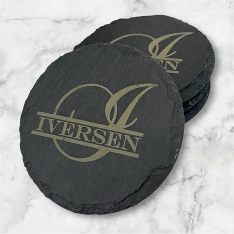 Enhance your space with the B. James Design & Woodwork Slate Coaster Set of 6—black round coasters customized with IVERSEN and a gold-engraved letter I, elegantly showcased on a white marble surface.