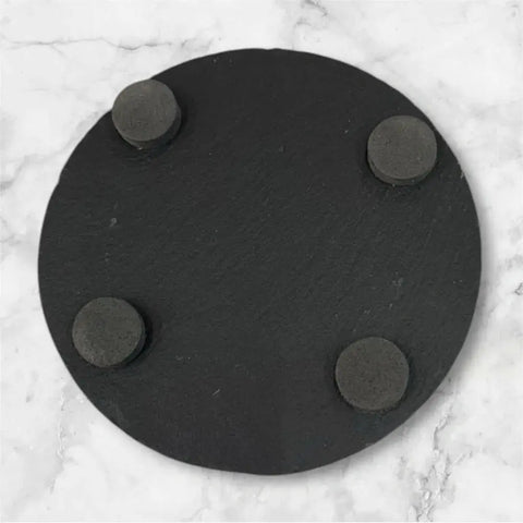 A round black slate coaster with four foam pads on the bottom, shown on a white marble surface—part of the Slate Coaster Set of 6 by B. James Design & Woodwork, ideal for custom personalization.