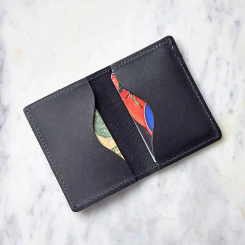 An Urban Southern Bifold Card Wallet in black rests open on a white marble surface, showing two colorful cards partially tucked into its slots.