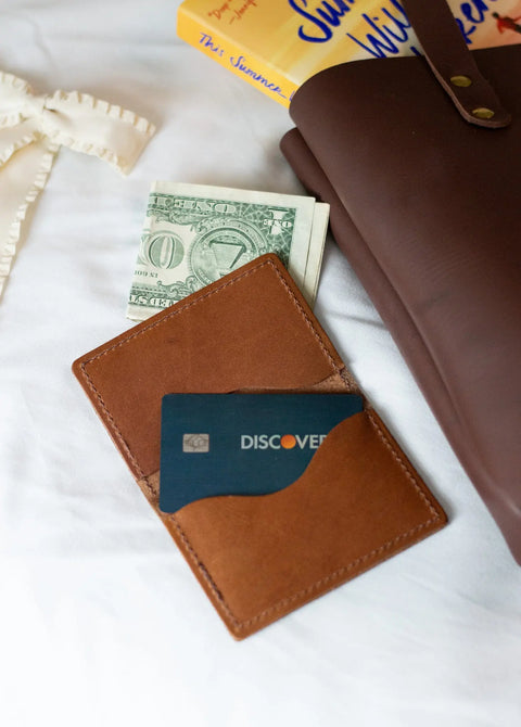 The Urban Southern Bifold Card Wallet, in brown, holds a Discover card and folded cash. It’s displayed on a white surface beside a brown notebook, a yellow book, and a cream ribbon.